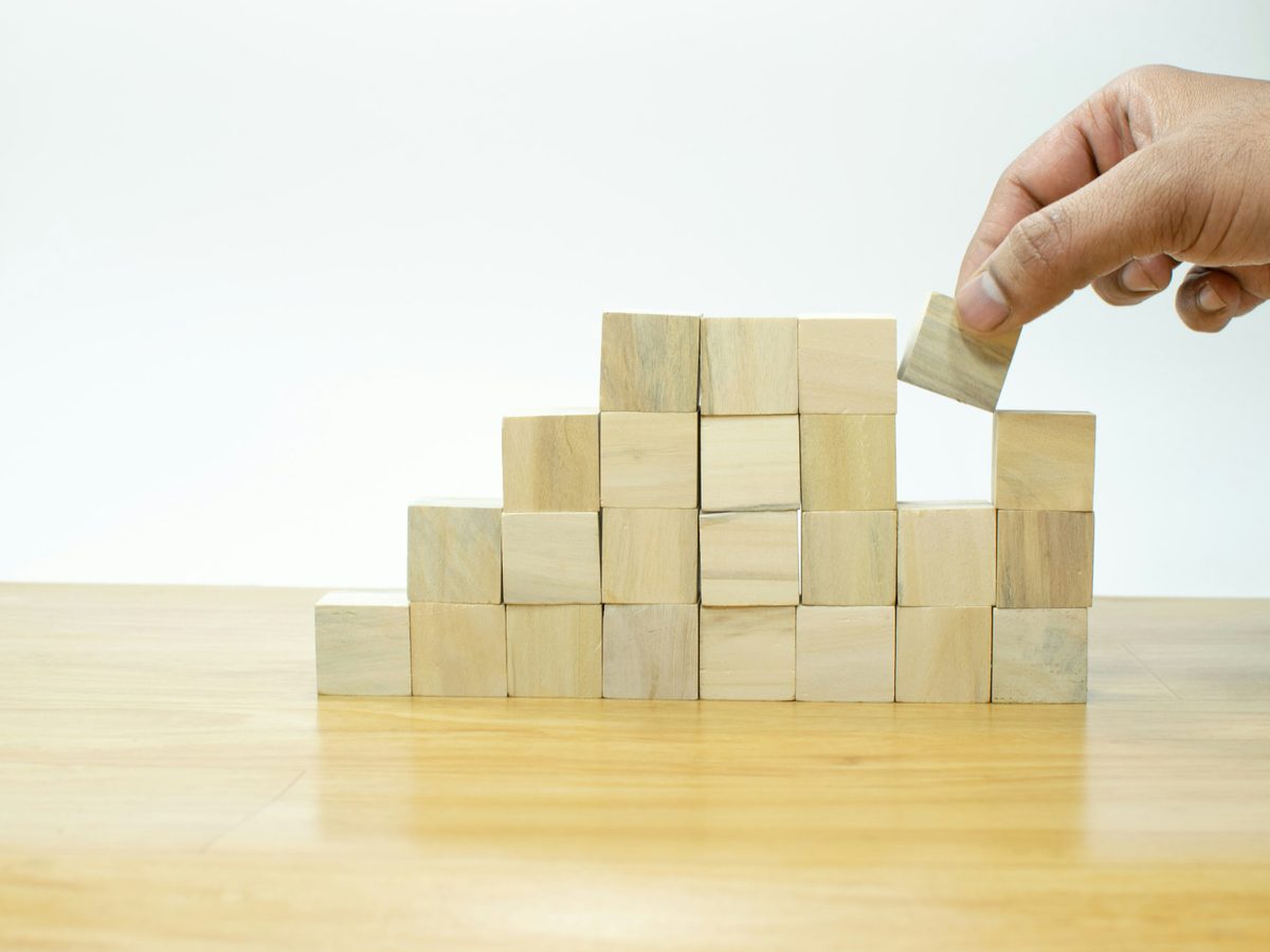 A hand placing the last wooden block on top of a pyramid of stacked blocks on a wooden desk.